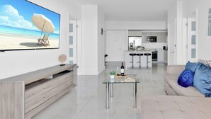 Un salon moderne avec un téléviseur à écran plat, une table basse en verre, un canapé et une cuisine en arrière-plan.