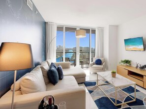 A modern living room with a sofa, a coffee table, a TV, and a view of a cityscape and water.