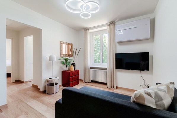 A modern living room with a flat-screen TV, a red cabinet, a floor lamp, and a window with curtains. - Cozy lake view apartment in Ruvigliana (DREAM LAKE 1) (Lugano)