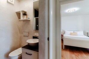 A compact bathroom with a toilet, sink, and storage cabinet. A bedroom with a bed and bedside table is visible through an open door. - Cozy lake view apartment in Ruvigliana (DREAM LAKE 1) (Lugano)