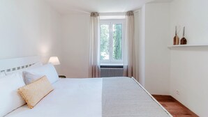 Un dormitorio con una cama, una mesita de noche, una ventana con cortinas y una estantería con dos jarrones.