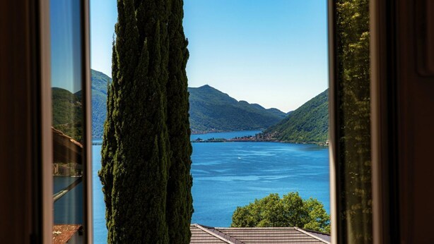 A view of a lake with mountains in the background, a tall tree in the foreground, and a building with a tiled roof. - Lovely one-bedroom apartment with balconies overlooking the lake (Dream Lake 3) (Lugano)