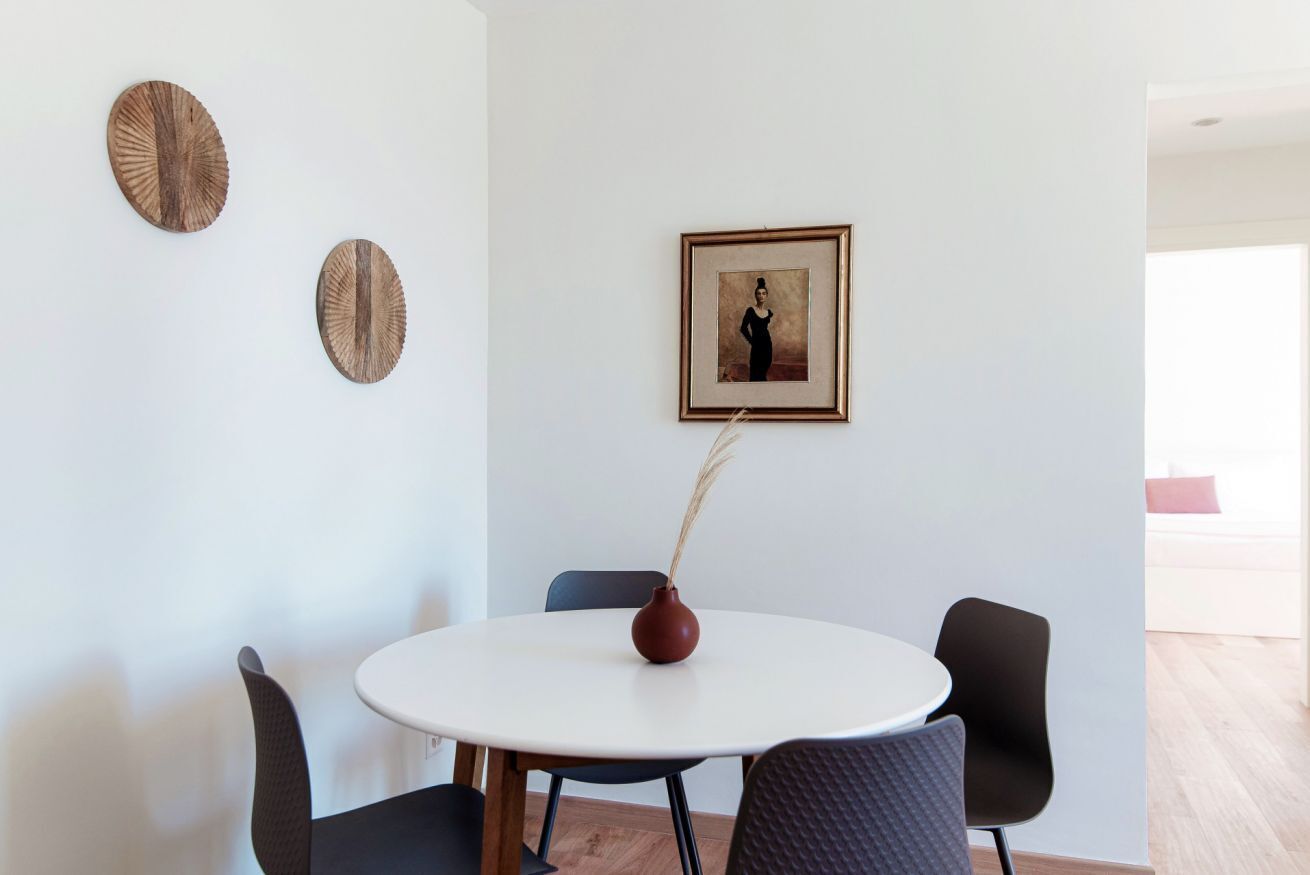 Et rundt spisebord med fire stole, en vase med tørrede planter, samt to dekorative genstande på væggen.
