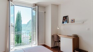 A bedroom with a bed, a desk, a chair, a small shelf with pictures, and a view of a mountain and lake through an open door. - Lovely one-bedroom apartment with balconies overlooking the lake (Dream Lake 3) (Lugano)