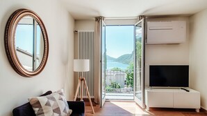 A modern living room with a sofa, a large mirror, a floor lamp, a TV on a white cabinet, and a sliding glass door leading to a balcony with a view of mountains. - Lovely one-bedroom apartment with balconies overlooking the lake (Dream Lake 3) (Lugano)