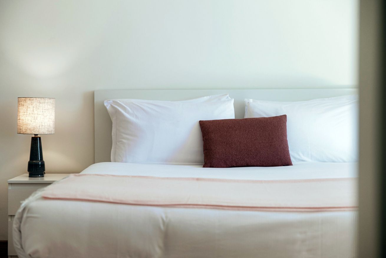 A neatly made bed with white linens and a single brown pillow, a bedside lamp, and a white nightstand.