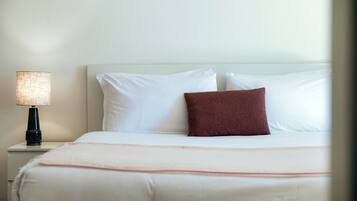 A neatly made bed with white linens and a single brown pillow, a bedside lamp, and a white nightstand.