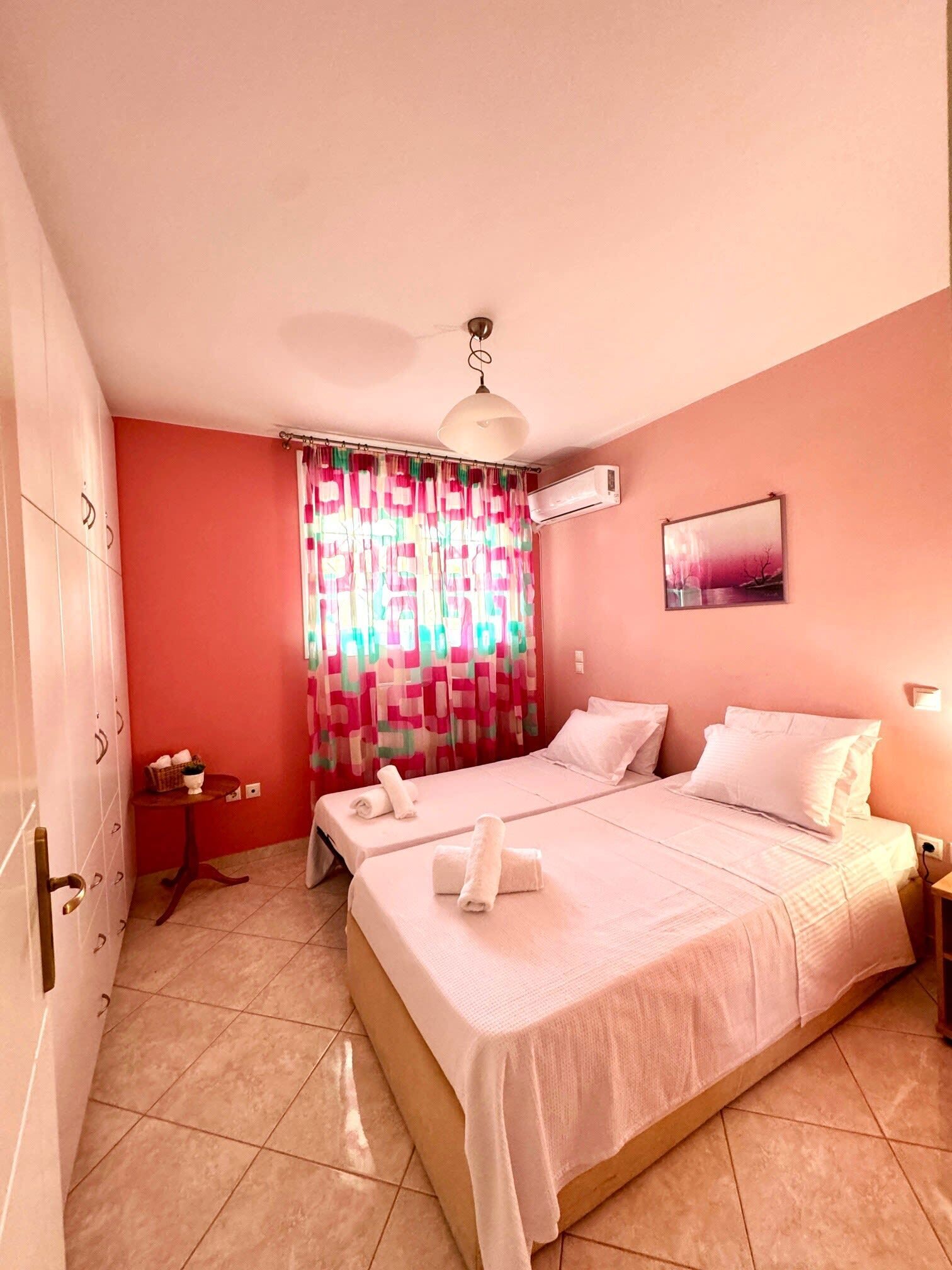 Une chambre d’hôtel avec un lit, une petite table sur laquelle se trouve un panier, un rideau, un appareil de climatisation et un tableau accroché au mur.