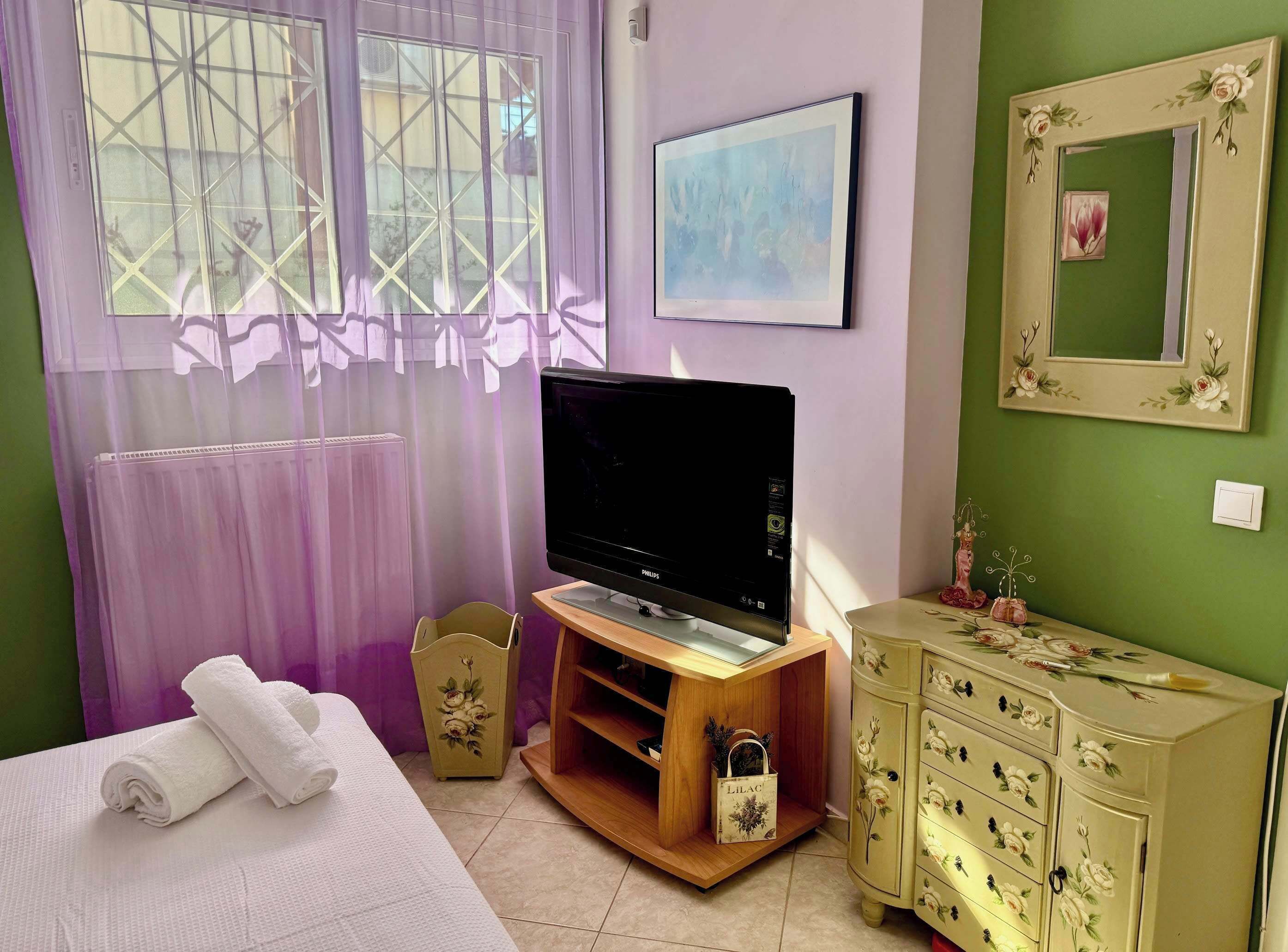 A room with a bed, a television on a wooden stand, a dresser, a window with curtains, and a painting on the wall.