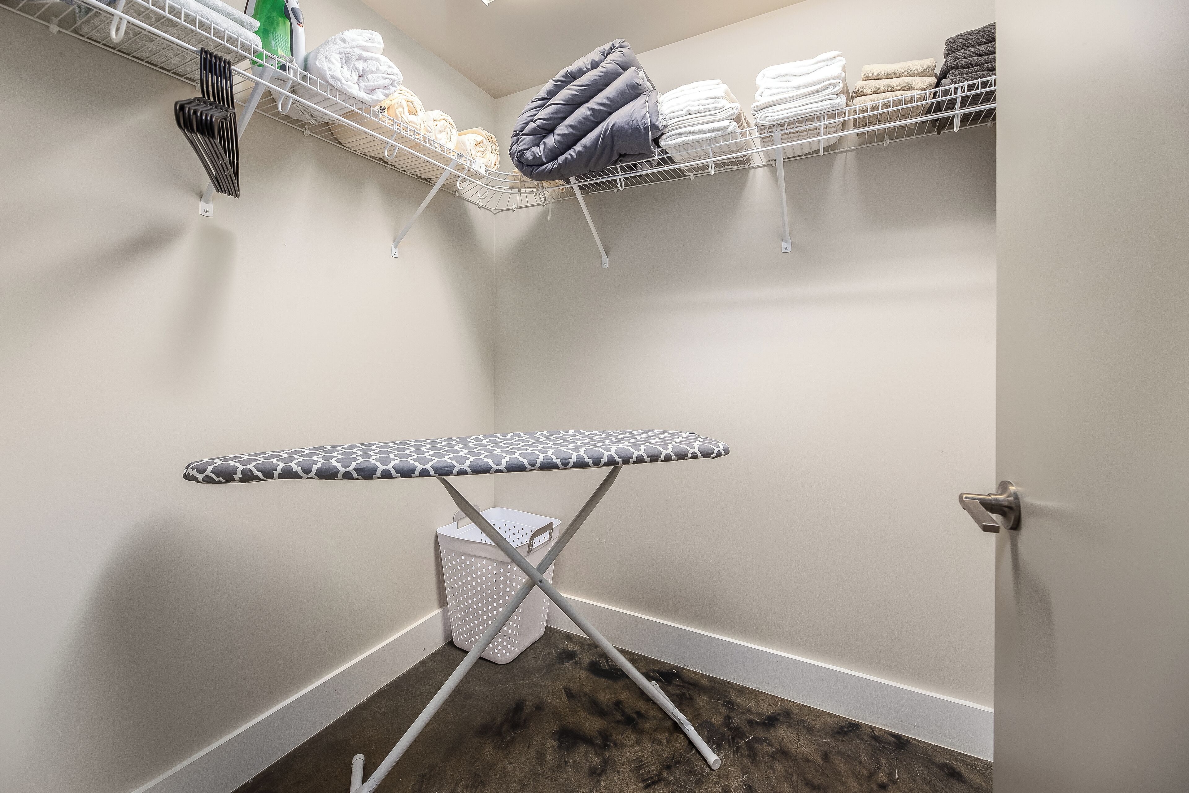 A corner laundry room with an ironing board, shelves with folded towels, and a laundry basket.