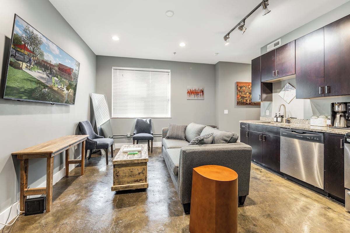 Un salon moderne avec un canapé gris, une table basse en bois et un fauteuil gris. Un grand tableau représentant un parc est accroché au mur, et la cuisine est équipée de meubles sombres et d’appareils électroménagers en acier inoxydable.
