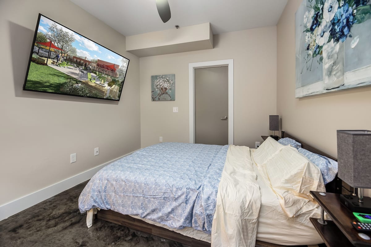 Une chambre à coucher avec un lit, un téléviseur fixé au mur, une porte et un tableau.