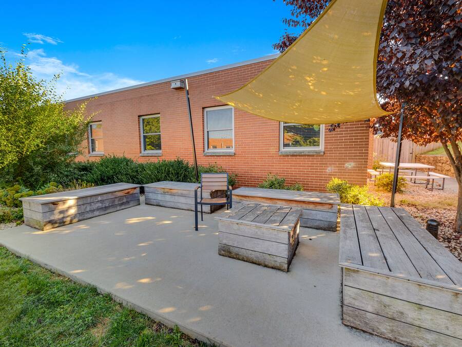 Un patio al aire libre con un banco y una mesa de madera, un edificio de ladrillo al fondo y una gran marquesina que proporciona sombra.