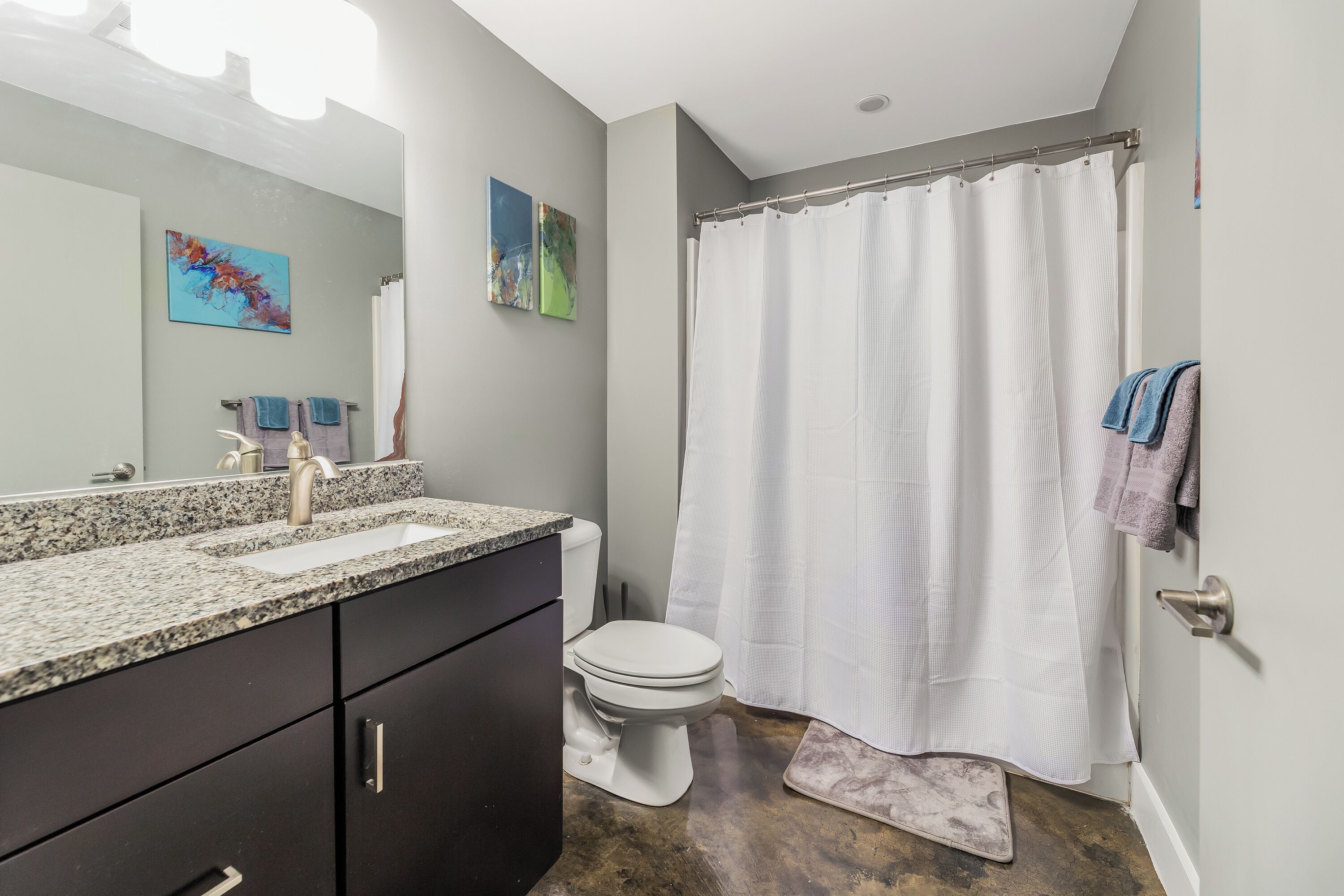 A modern bathroom with a granite countertop, a white toilet, a shower curtain, and a small rug.