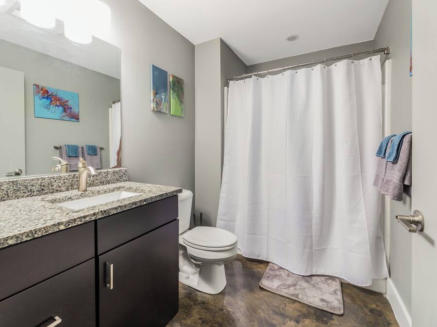 A modern bathroom with a granite countertop, a white toilet, a shower curtain, and a small rug.