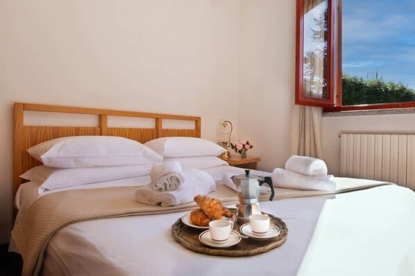 A neatly made bed with white linens, a tray with croissants and coffee cups, and a view of greenery through a window.