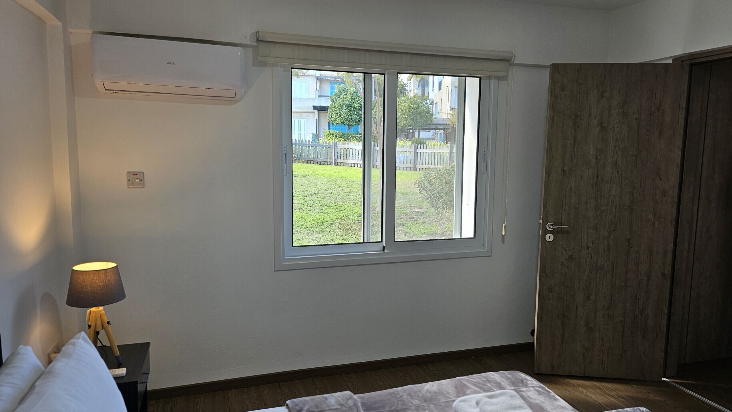 A bedroom with a window, a bedside lamp, a wooden door, and a wall-mounted air conditioner.