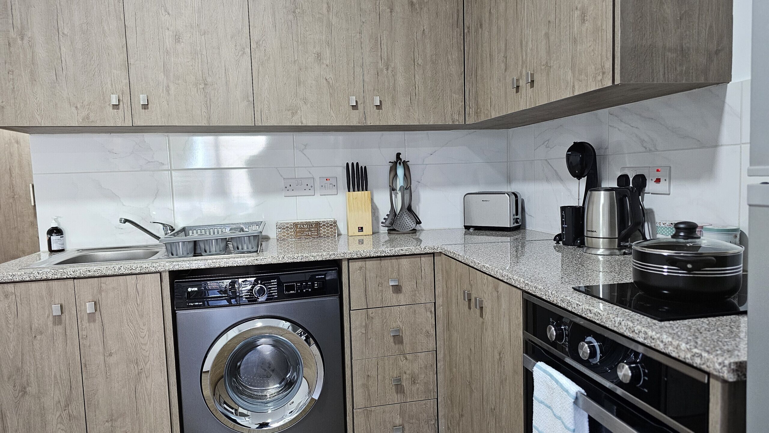 A compact kitchen with wooden cabinets, a washing machine, a toaster, and a coffee maker.