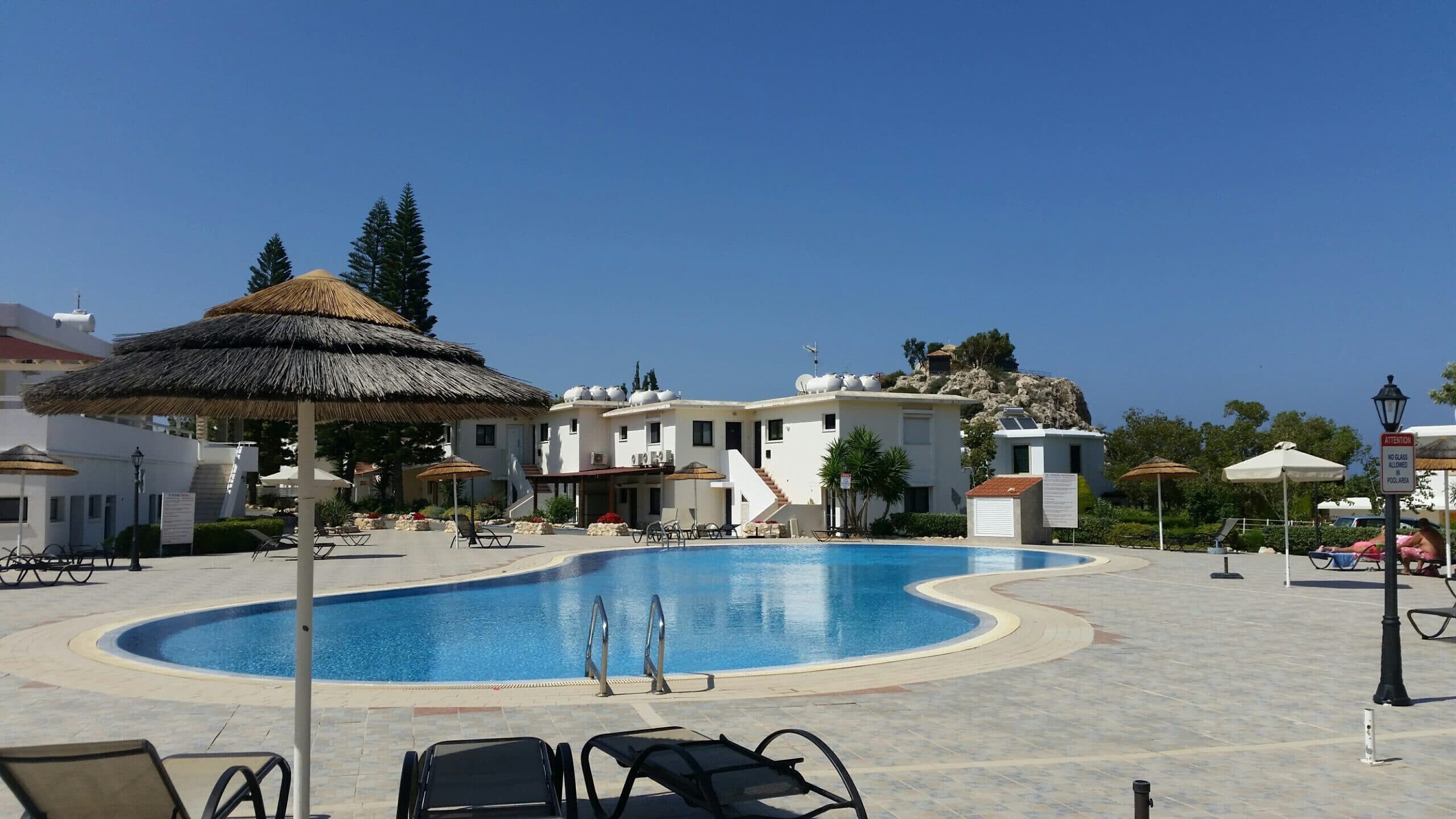A swimming pool with lounge chairs and umbrellas, surrounded by buildings with a mountainous backdrop.
