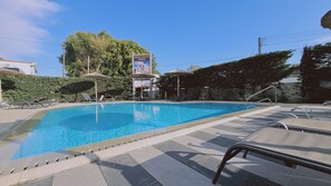 Una piscina con sillones de descanso y sombrillas, rodeada de vegetación y un cielo despejado.