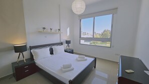 A modern bedroom with a large bed, bedside tables, a desk, a window with a view, and a hanging light fixture. - STAY White Petals Apartment (Larnaca)
