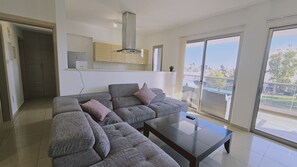 A modern living room with a grey sectional sofa, a glass coffee table, and a view of the outdoors through large windows. - STAY White Petals Apartment (Larnaca)