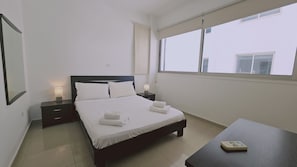 A hotel room with a bed, bedside tables, a window, and a small table. - STAY White Petals Apartment (Larnaca)