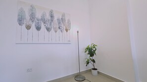 A room with a white wall, a floor lamp, a potted plant, and a painting of abstract tree-like structures. - STAY White Petals Apartment (Larnaca)