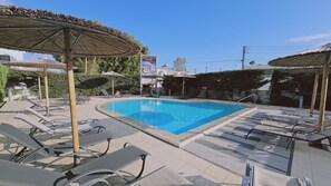 A swimming pool with lounge chairs and umbrellas, surrounded by greenery and a clear sky. - STAY White Petals Apartment (Larnaca)