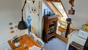 Ein gemütliches Zimmer mit einem Holzesstisch, weißen Stühlen, einem kleinen weißen Kühlschrank und einer Erkerzone mit einer Holzleiter.