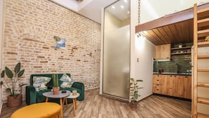 Un espace de vie moderne avec un mur en briques, un canapé vert, un pouf jaune et un coin cuisine doté de meubles en bois et d’une étagère suspendue.
