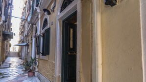 Une ruelle étroite pavée de pierres, bordée de bâtiments des deux côtés, avec une porte noire et des plantes en pot.