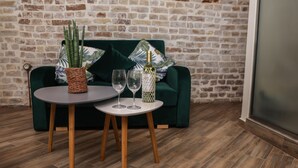 Une pièce avec un mur en briques, un canapé vert orné de coussins à motifs, une table ronde avec deux verres à vin et une bouteille, et un pot à plantes en osier.