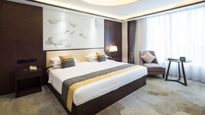 Habitación de hotel con una cama grande, una mesita de noche, una silla, una pequeña mesa y un ventanal con cortinas.