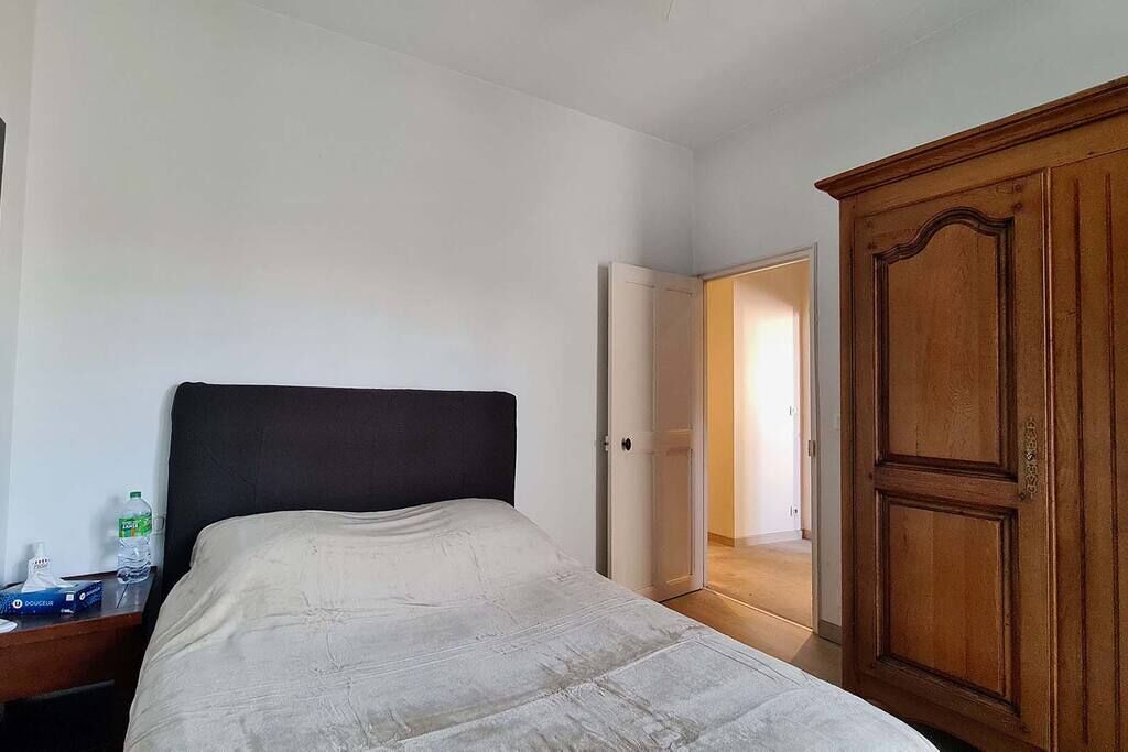 A hotel room with a bed, a wooden wardrobe, a nightstand with a bottle, and a door leading to another room.