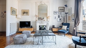 A living room with a fireplace, framed pictures, a television, and a blue armchair. - Clichy Couture, by Veeve (Paris)