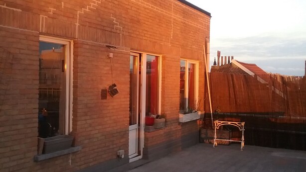 Een gebouw van baksteen met grote ramen en een deur, een klein buitentafeltje, en een heldere lucht.
