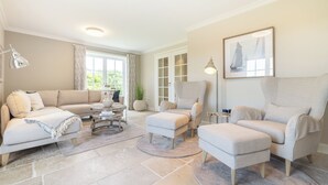 Un salon spacieux avec un canapé beige, un grand fauteuil et une table basse. Il y a une grande fenêtre avec des rideaux, un tableau au mur et une plante en pot.