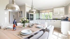 Une cuisine moderne avec des meubles blancs, une table à manger en bois pour six personnes et des luminaires suspendus.