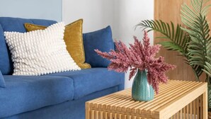 Een blauwe bank met een wit en goud kussen, een houten bijzettafeltje met een vaas vol roze bloemen, en een groene plant.