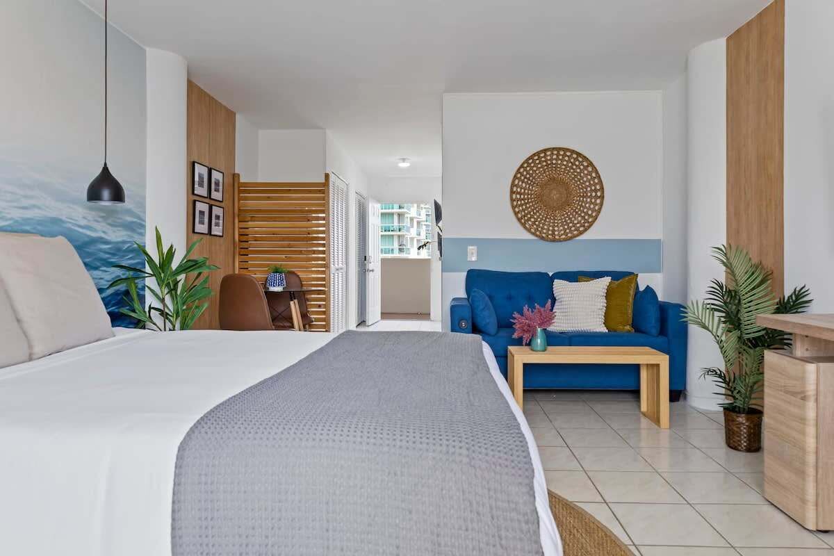 A modern hotel room with a large bed, a blue sofa, a wooden side table, and a wall-mounted decorative item.