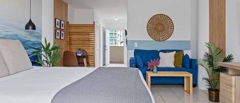 A modern hotel room with a large bed, a blue sofa, a wooden side table, and a wall-mounted decorative item.