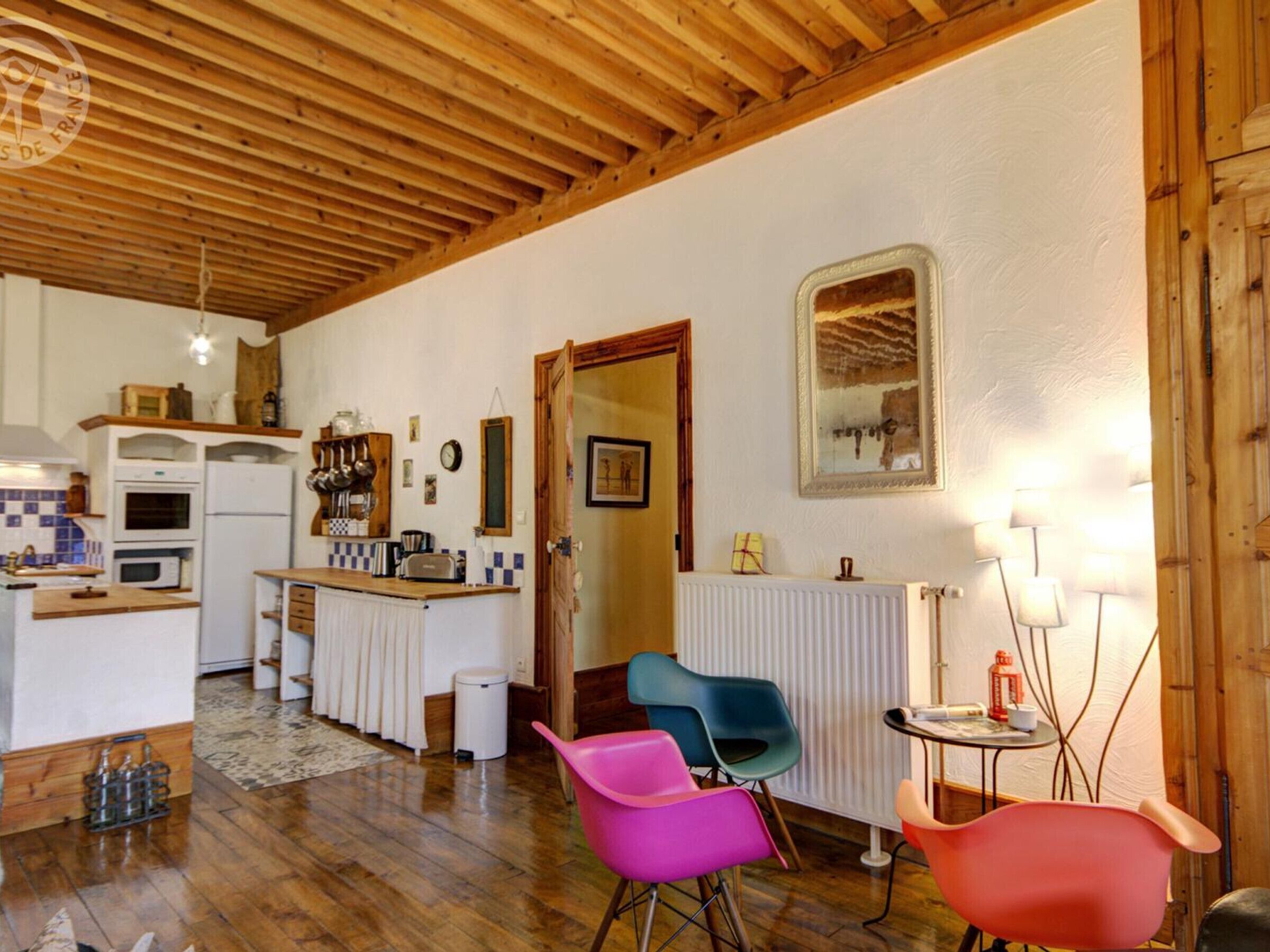 A cozy kitchen with wooden beams, a white fridge, and a small dining area with colorful chairs.
