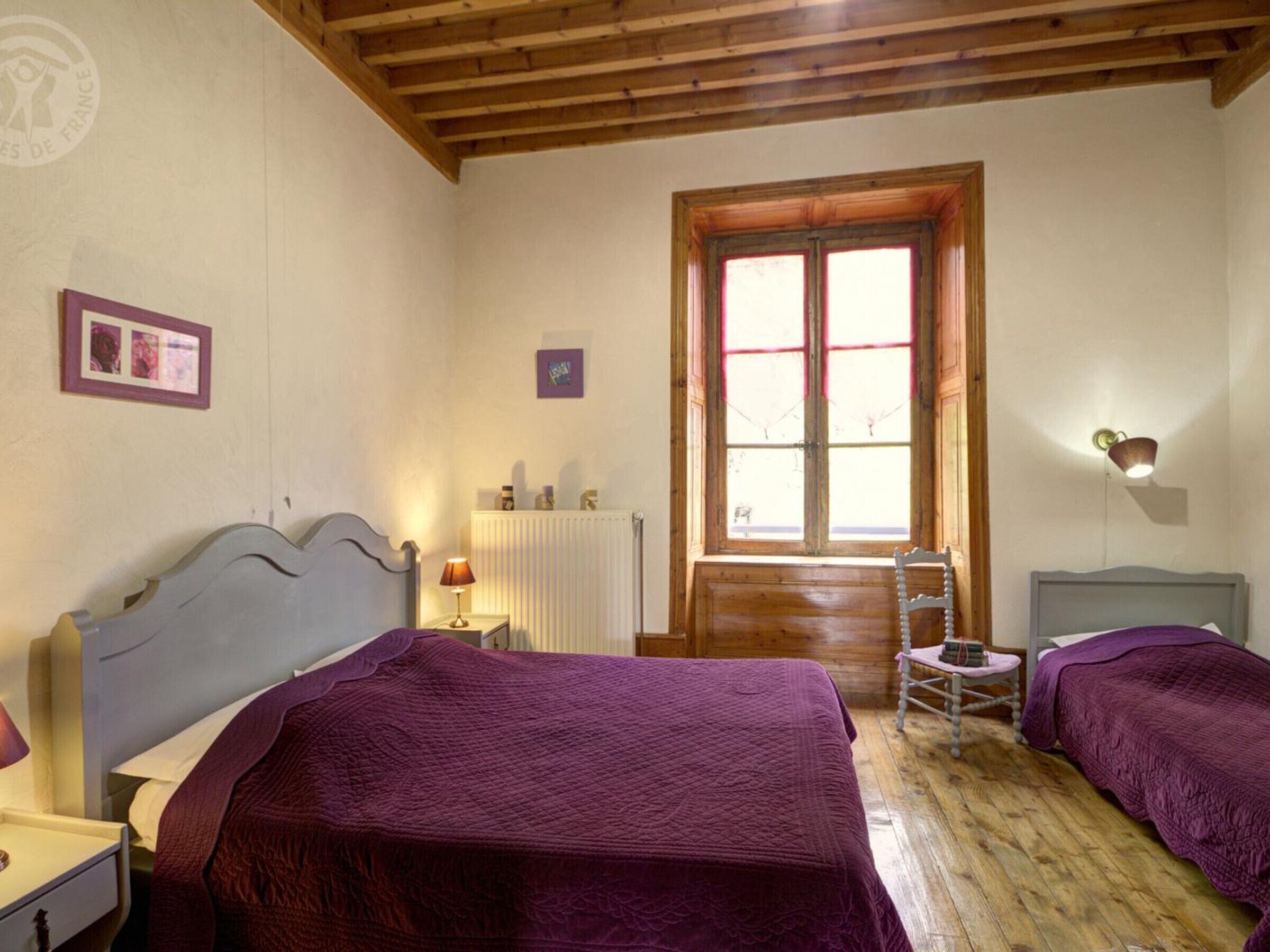 A room with two beds, wooden beams on the ceiling, a window with shutters, a radiator, and a small table with a chair.