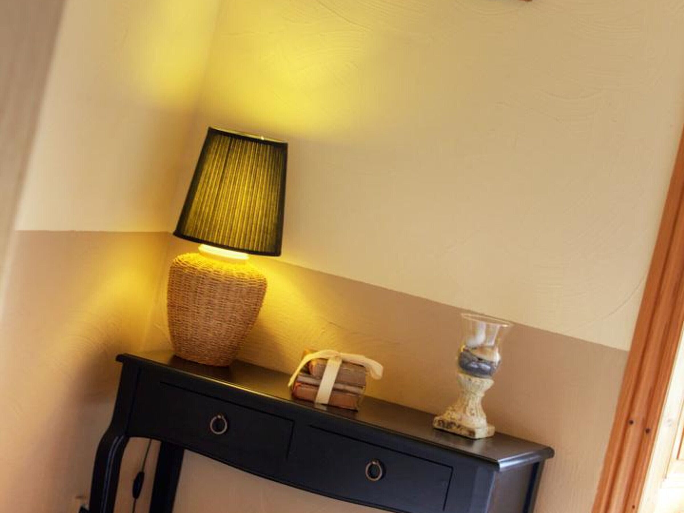 Une petite console de couleur sombre, agrémentée d’un panier tressé, d’une lampe avec un abat-jour vert et d’un vase décoratif.
