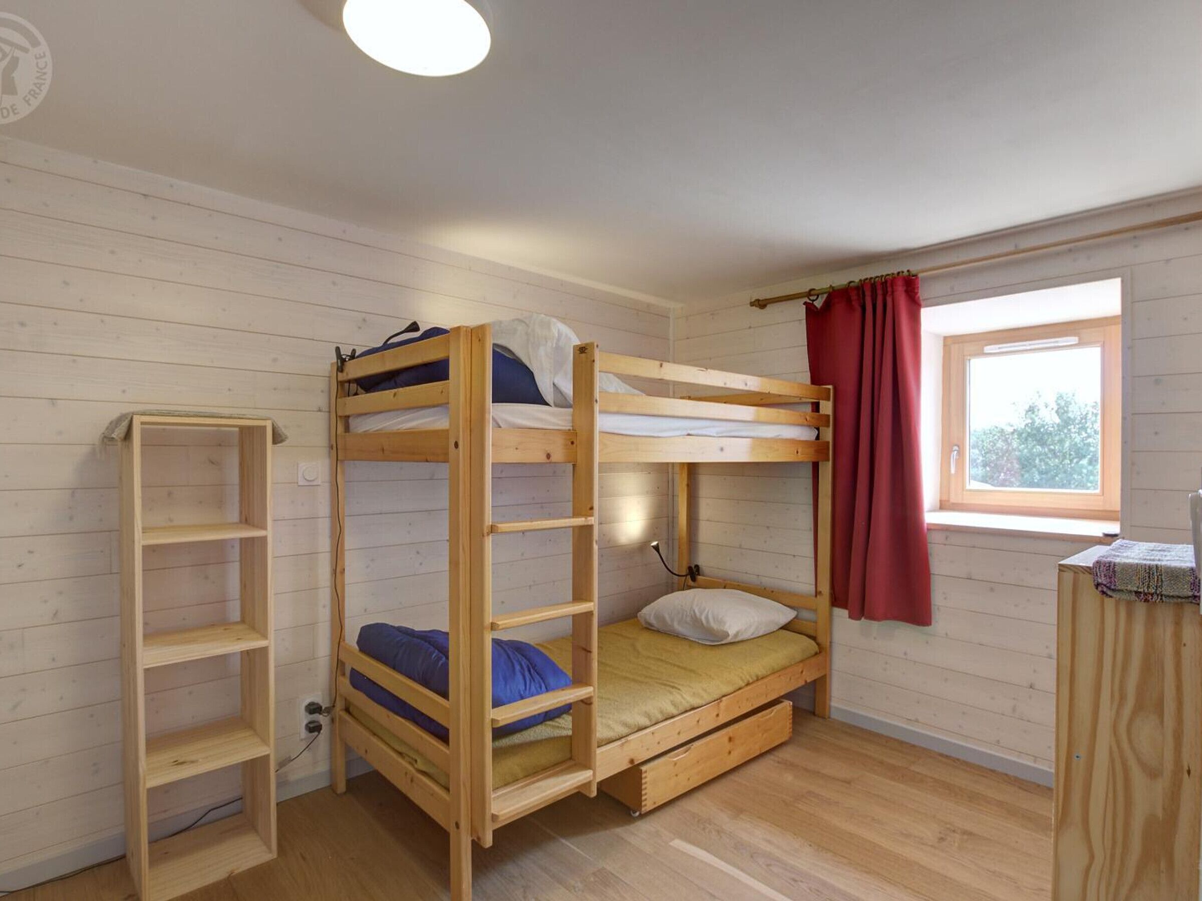 Habitación con literas, una escalera de madera, un escritorio pequeño y una ventana con cortinas rojas.