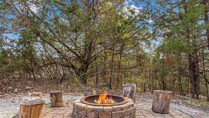 A circular stone fire pit with a fire, surrounded by wooden stumps, set in a paved area with trees in the background. - Lakeview! Hot Tub & Fire pit! Game Room! (Hollister)