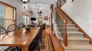 A spacious dining area with a long wooden table, chairs, and a chandelier. A staircase with wooden railings leads to an upper level. - Lakeview! Hot Tub & Fire pit! Game Room! (Hollister)