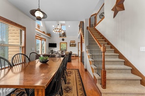 Un comedor amplio con una larga mesa de madera, sillas y una araña. Una escalera con barandillas de madera conduce a un nivel superior.