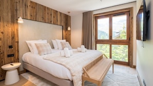 Ein modernes Schlafzimmer mit einem Holz-Kopfteil, einem großen Bett mit weißen Bettwäsche, einer Holzbank, einem Nachttisch und einem großen Fenster mit Blick auf Grünflächen.